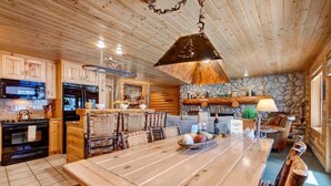A rustic cabin interior with a wooden dining table, kitchen area, stone fireplace, and log walls.