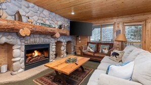 A cozy cabin interior with a stone fireplace, wooden ceiling, a leather sofa, and a television mounted on the wall.