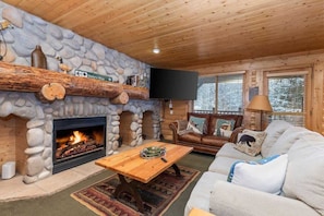 A cozy cabin interior with a stone fireplace, wooden ceiling, a leather sofa, and a television mounted on the wall.