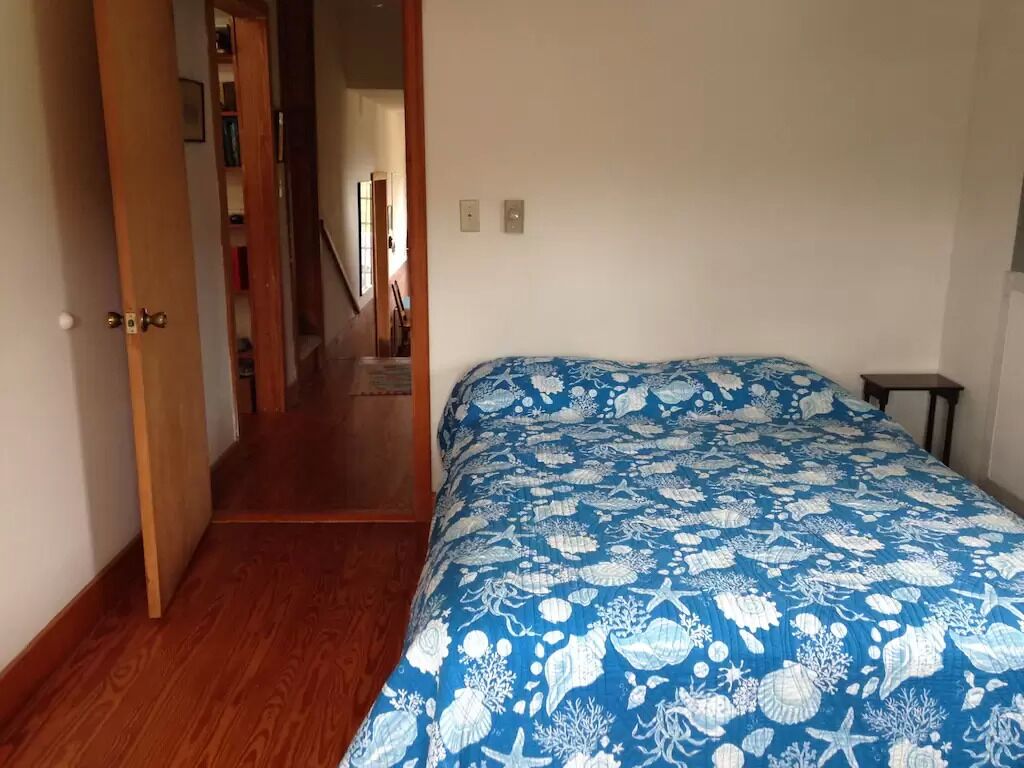 A bedroom with a bed, wooden flooring, a door leading to a hallway, and a small table.