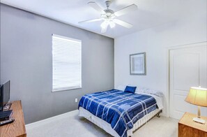 Une chambre à coucher avec un lit, un ventilateur de plafond, un bureau avec un ordinateur et une lampe.