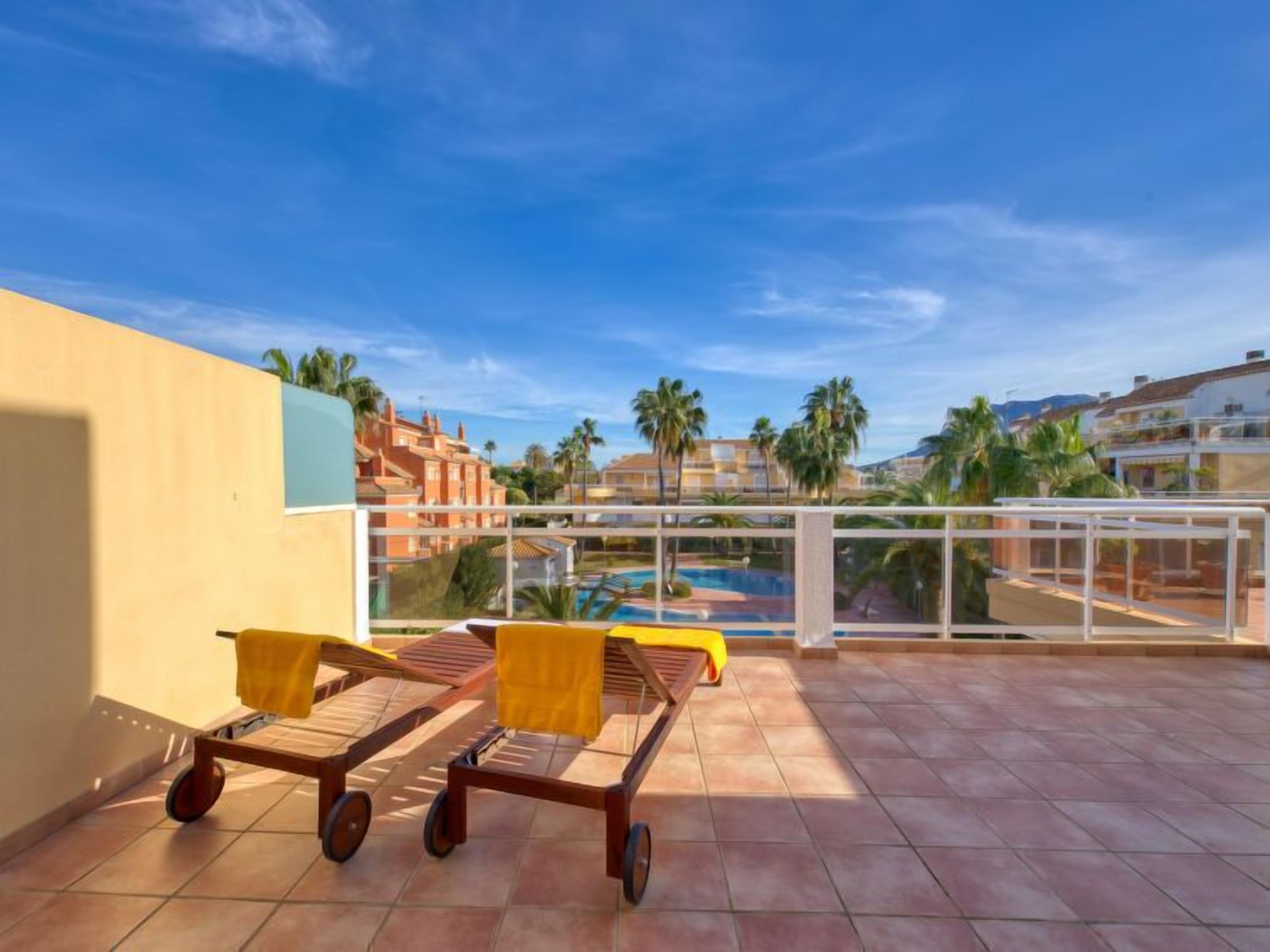 A rooftop terrace with two lounge chairs, a pool, and a view of palm trees and buildings.