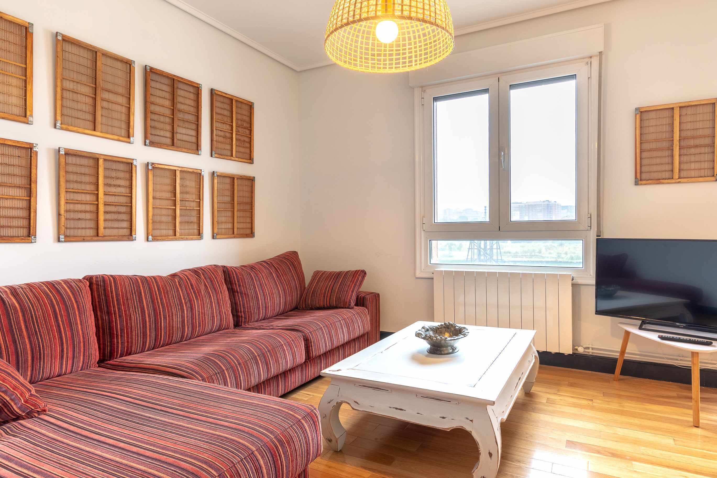 Un salón con un sofá a rayas, una mesa de centro blanca, un televisor y una ventana con persianas.
