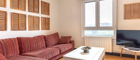 Un salón con un sofá a rayas, una mesa de centro blanca, un televisor y una ventana con persianas.