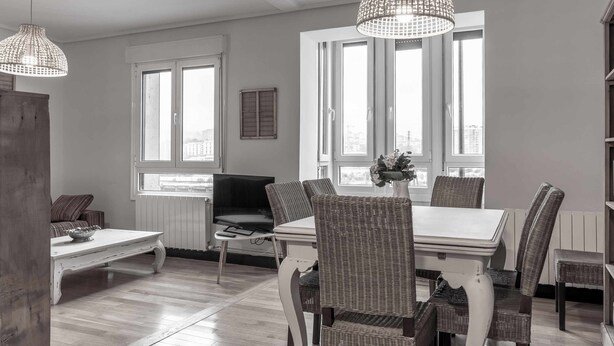 A dining area with a white table and wicker chairs, large windows with blinds, and a hanging light fixture.