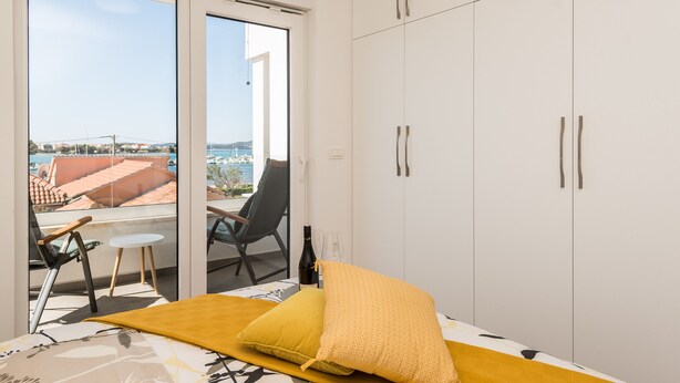 A bedroom with a bed, a yellow pillow, a bottle of wine, and a view of the sea through a sliding glass door. - Apartment Birgir in Sukošan (Sukošan)