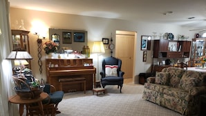 A living room with a piano, floral sofa, and a chair. There is a wooden cabinet with decorative items and a lamp.