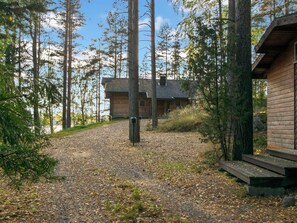 A cabin with a gravel driveway, surrounded by tall trees and a clear sky.