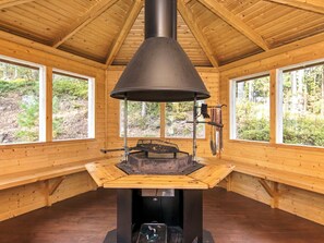 A cabin interior with a central wood-burning stove, large windows, and a view of the forest.