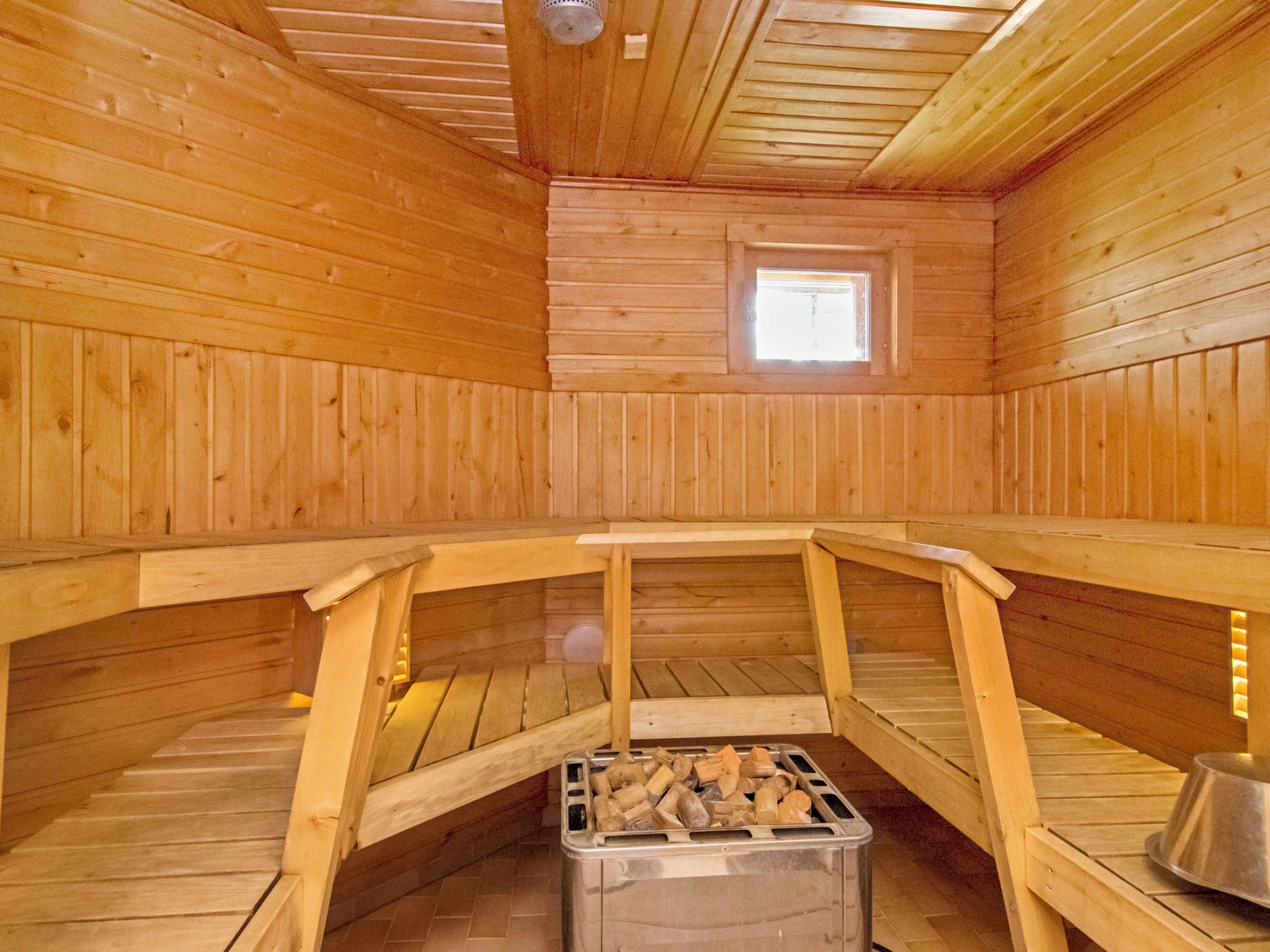 A sauna with wooden walls and benches, a skylight, and a metal container with logs.