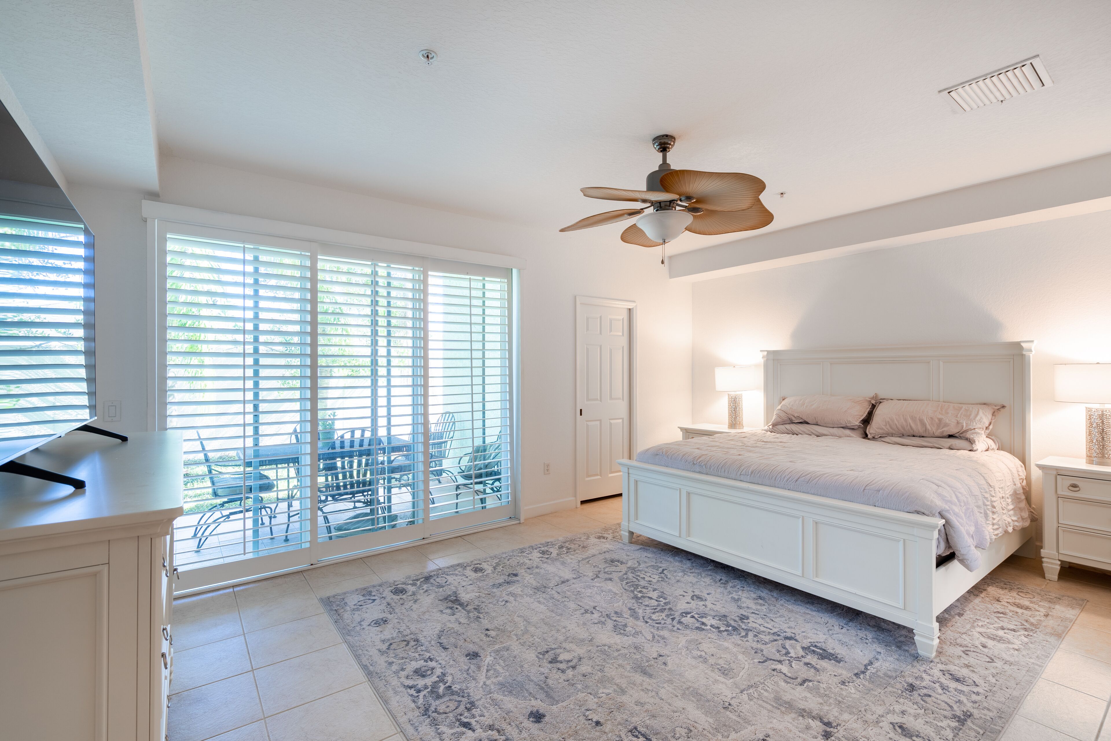 A bedroom with a large bed, a ceiling fan, a sliding glass door leading to a balcony, and a small desk with a lamp.