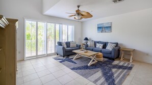 Un salon spacieux avec un plafond haut, un ventilateur de plafond, une grande fenêtre avec des stores, un ensemble de canapés bleus, une table basse et un tableau accroché au mur.