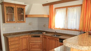 A kitchen with wooden cabinets, a granite countertop, a built-in dishwasher, and a window with orange curtains. - Haus Falke by Interhome (Saas-Grund)