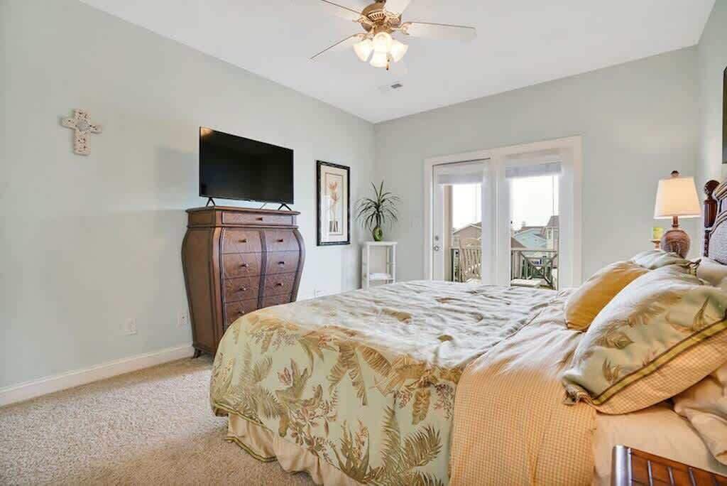 Une chambre à coucher comprenant un lit, une télévision, un ventilateur de plafond, une croix accrochée au mur et une porte donnant sur un balcon.