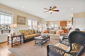 A spacious living room with a ceiling fan, two sofas, a coffee table, and a dining area with wooden cabinets. - Doin` Time by Brunswick Vacation Rentals: 5BR Ocean View Home w/ Dock & Elevator (Ocean Isle Beach)