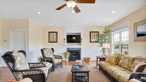 A living room with wicker furniture, a fireplace, a ceiling fan, and framed artwork on the walls. - Doin` Time by Brunswick Vacation Rentals: 5BR Ocean View Home w/ Dock & Elevator (Ocean Isle Beach)