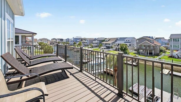 A deck with lounge chairs overlooking a waterway and neighboring houses. - Doin` Time by Brunswick Vacation Rentals: 5BR Ocean View Home w/ Dock & Elevator (Ocean Isle Beach)