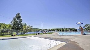 A swimming pool area with a water slide, a lifeguard tower, and a fenced area with trees and a lake in the background.