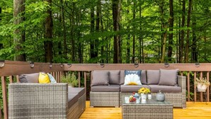 An outdoor deck with wicker furniture and a coffee table, surrounded by a wooden railing and a forest backdrop.