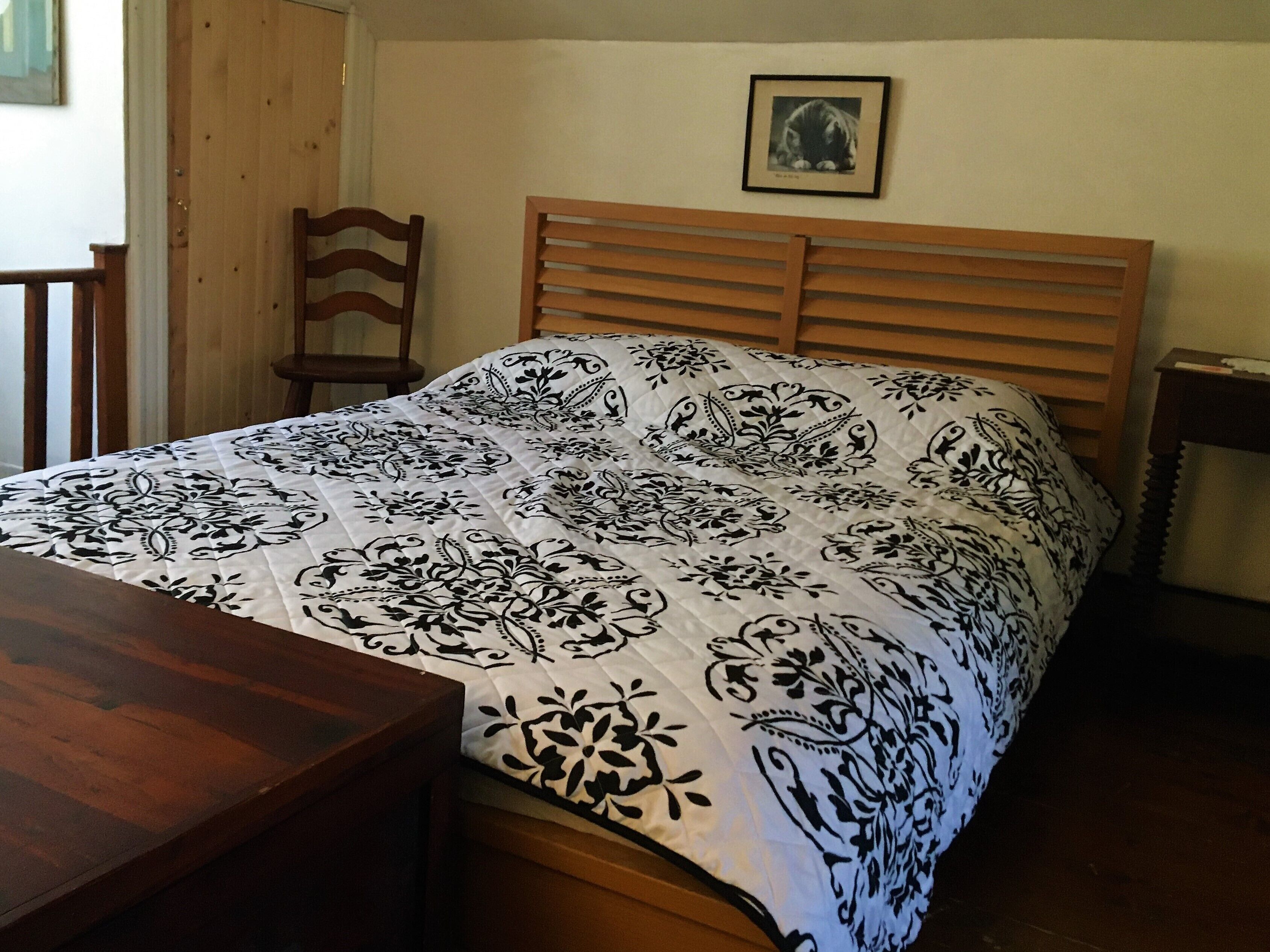 A bedroom with a wooden bed frame, a white and black patterned bedspread, a wooden nightstand, and a framed picture on the wall.