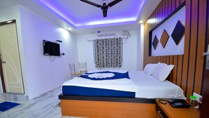 Quarto de hotel com cama, ventoinha no teto, televisão e mesa de cabeceira com telefone e uma xícara.