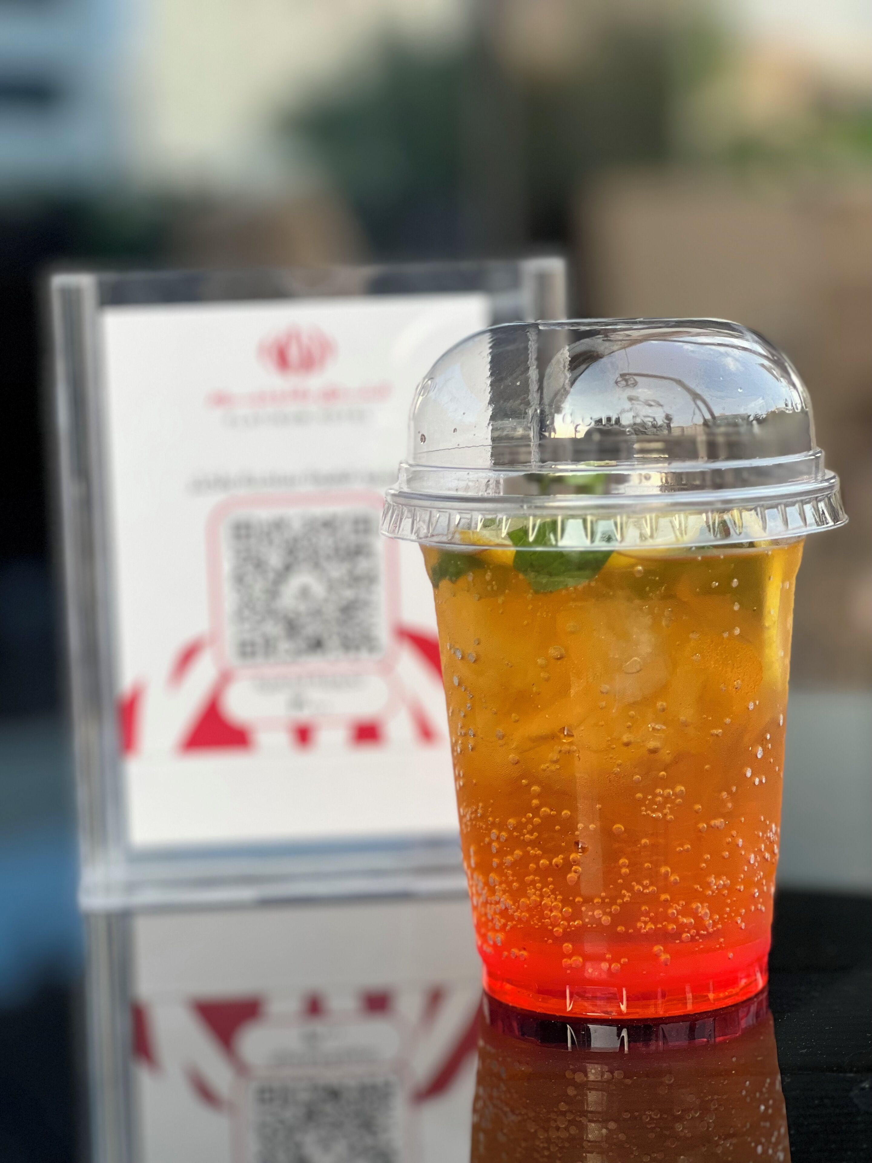 A clear plastic cup with a red bottom and a transparent top, filled with a yellow-orange beverage and ice cubes, placed on a reflective surface.