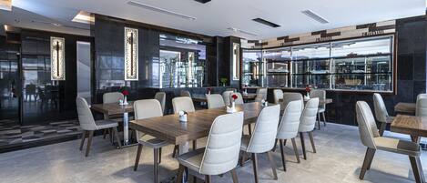 A modern restaurant with a long wooden table and white cushioned chairs, a glass display case, and a black and white color scheme.
