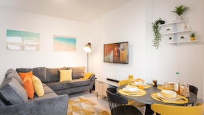 Un salon moderne avec un canapé gris, des coussins jaunes, une table à manger avec des chaises jaunes et un téléviseur fixé au mur.