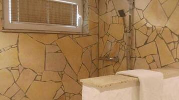 A bathroom with a stone wall, a shower, and a window with blinds.