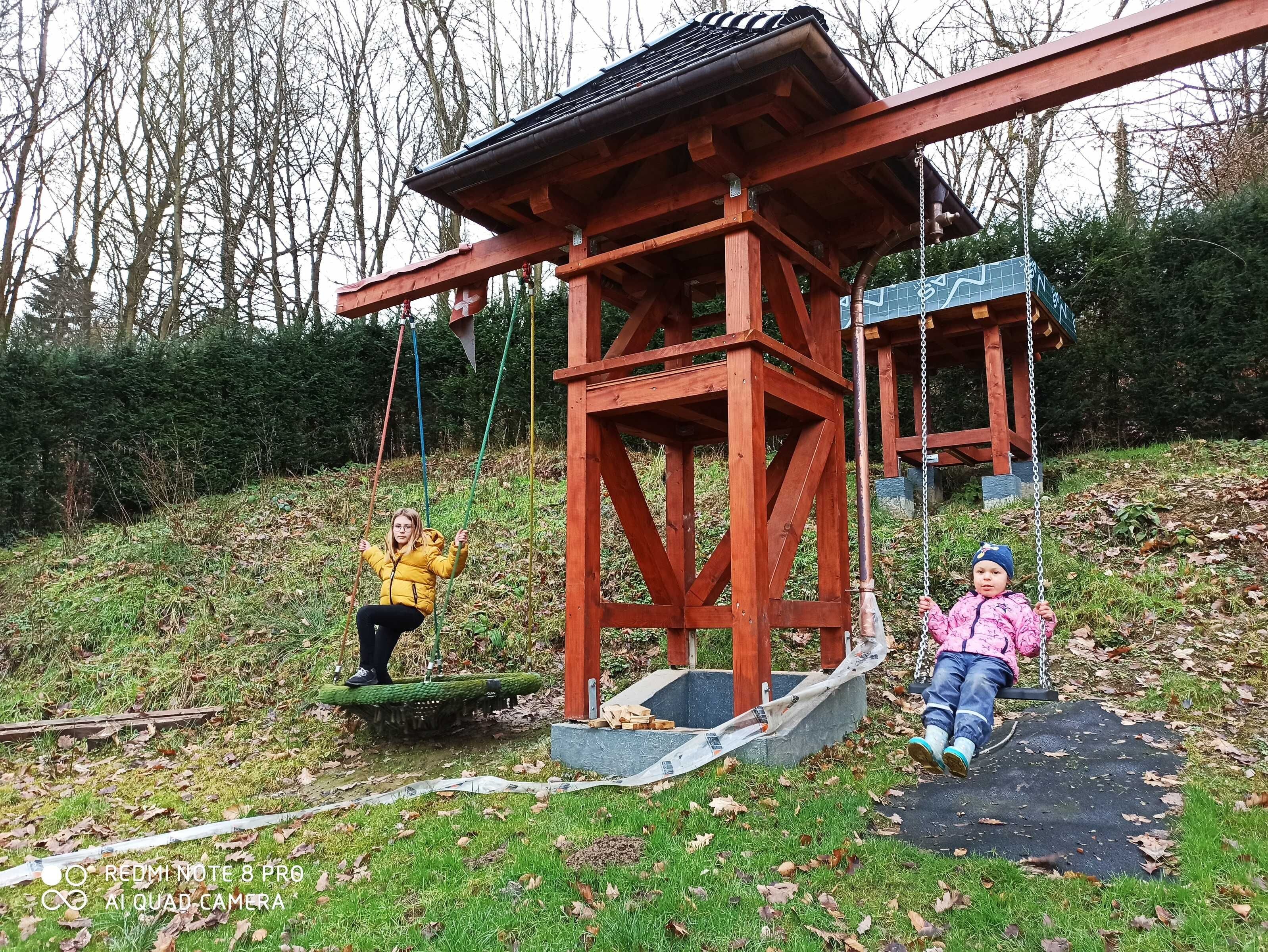 A wooden swing set with a metal frame, a small covered structure, and a grassy area with trees in the background.
