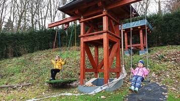 A wooden swing set with a metal frame, a small covered structure, and a grassy area with trees in the background.