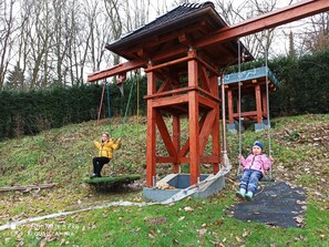 Un conjunto de columpios de madera con estructura metálica, un pequeño refugio cubierto y una zona con césped y árboles al fondo.