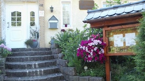 Un edificio con una puerta blanca, una escalera de piedra que conduce a una estructura de madera y un jardín con plantas en flor.