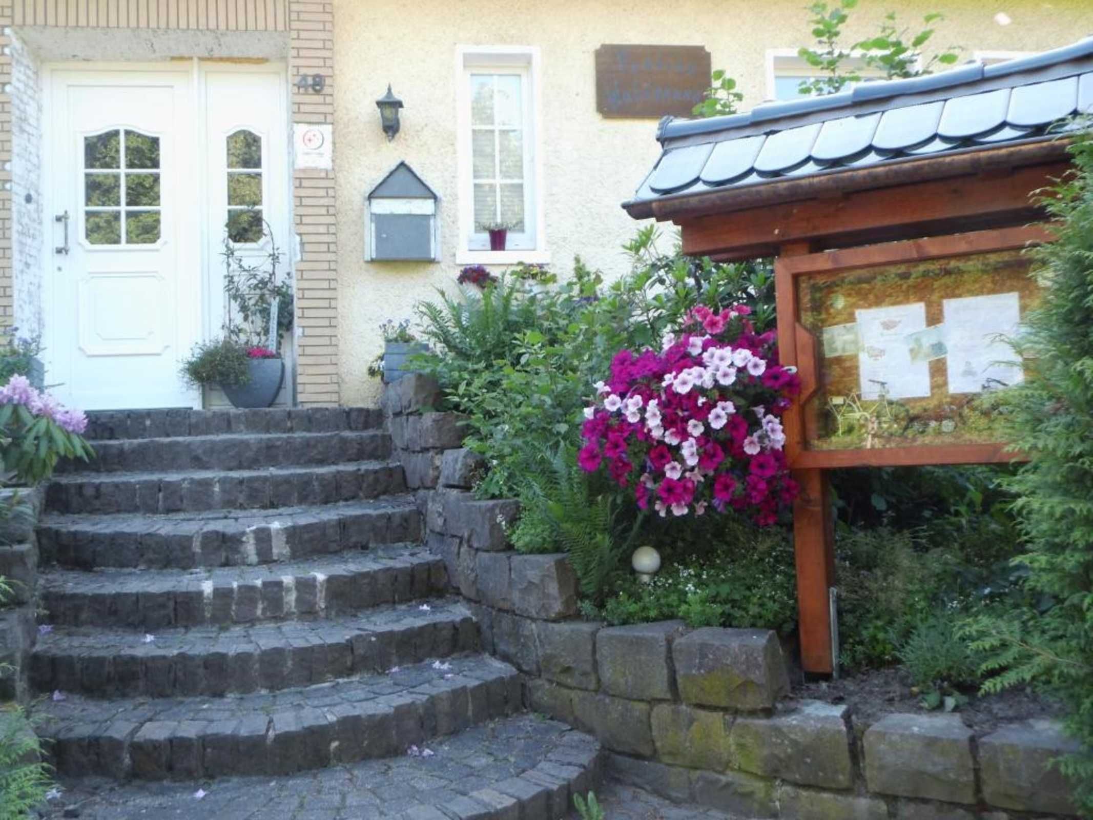 Ein Gebäude mit einer weißen Tür, einer Treppe aus Stein, die zu einer Holzkonstruktion führt, und einem Garten mit blühenden Pflanzen.