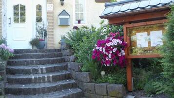 A building with a white door, a set of stone steps leading to a wooden structure, and a garden with flowering plants.