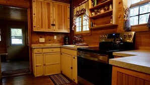 A wooden cabin interior with a kitchen area featuring wooden cabinets, a stove, and a window with curtains.