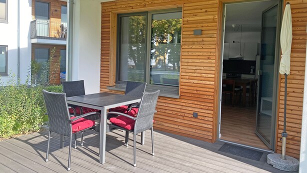 Een houten terras met een tafel en stoelen, een schuifdeur naar een keuken en een uitzicht op een woongebouw.
