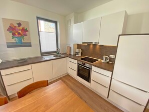Une cuisine moderne avec des meubles blancs, un four encastré, un évier et une fenêtre équipée de stores.
