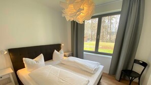 Een tweepersoonsbed met witte lakens, een donker hoofdbord, een nachtkastje met een lamp, en uitzicht op een grasveld door een raam met gordijnen.