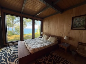 A bedroom with a large bed, wooden walls, a painting, and a view of the outdoors. - Feeling Superior - Cliffside Home with Amazing Lake Views (La Pointe)