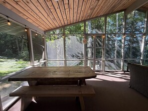 Ein überdachter Außenbereich mit Holztisch und -bänken, großen Fenstern und Blick auf den Wald.