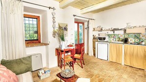 A cozy living room with a sofa, a small table with a book, and a window with curtains. Adjacent is a kitchen with a microwave, a sink, and a tiled backsplash.