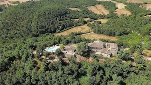 An aerial view of a rural property with a stone house surrounded by dense forest, a swimming pool, and terraced fields.