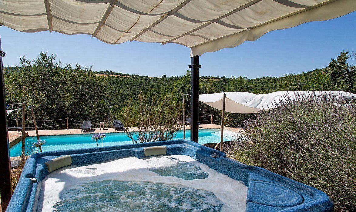 A hot tub with a pool in the background, surrounded by trees and a clear sky.