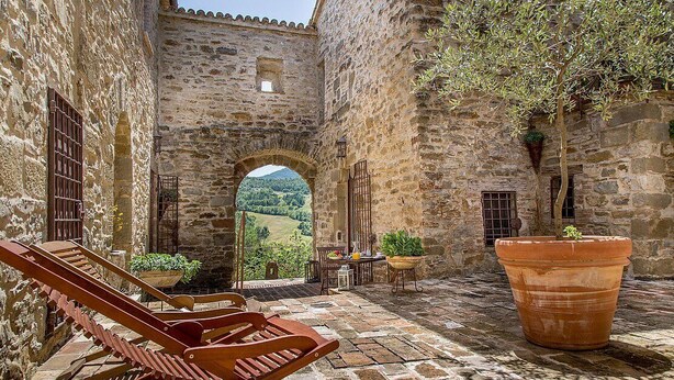 A stone courtyard with a view of the countryside, wooden chairs, a table, and a large terracotta pot.