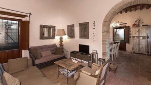 A cozy living room with a sofa, a coffee table, and a TV. There is a fireplace, a window with a view, and a dining area with chairs and a table.