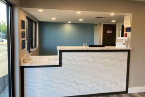 A reception desk with a marble top and a textured white panel. There are signs on the wall and a door with a fire safety sign. - Homelodge Newnan (Newnan)