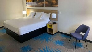 A hotel room with a large bed, a nightstand with a lamp, a chair, and a wall-mounted artwork. - Homelodge Newnan (Newnan)