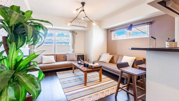 Een moderne woonkamer met een bank, een salontafel en een eetgedeelte met stoelen. In de hoek staat een grote plant en er is een raam met uitzicht.