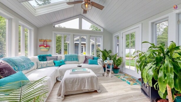 Una stanza luminosa con soffitto bianco, ampie finestre, un divano bianco con cuscini color acquamarina e un tavolino.